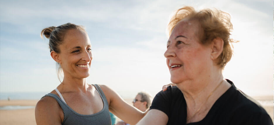 Lächelnde Physiotherapeutin, die einer älteren Frau bei Dehnübungen am Strand hilft.
