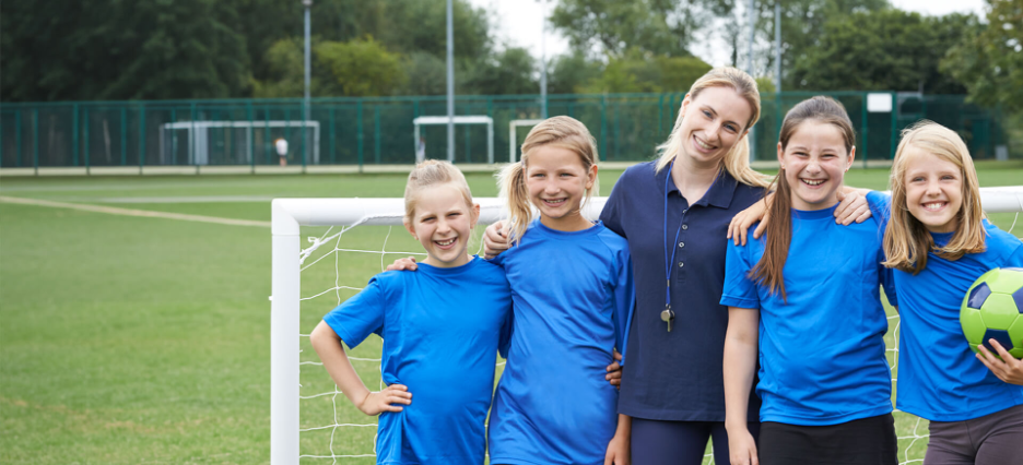 Sportsozialarbeiterin legt ihre Arme um eine Gruppe junger Fußballerinnen.